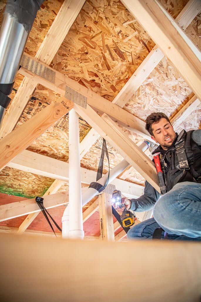 Two people install construction in an unfinished attic space, using power tools.