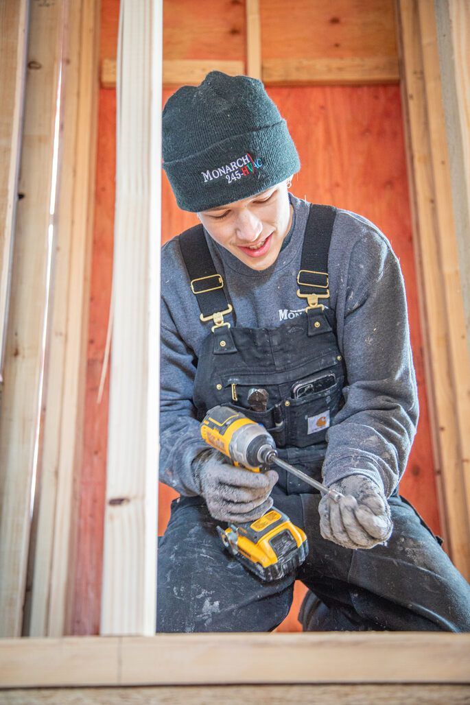 Person kneels, using a drill on wood framing. Wearing overalls, gloves, and a hat. Construction setting.
