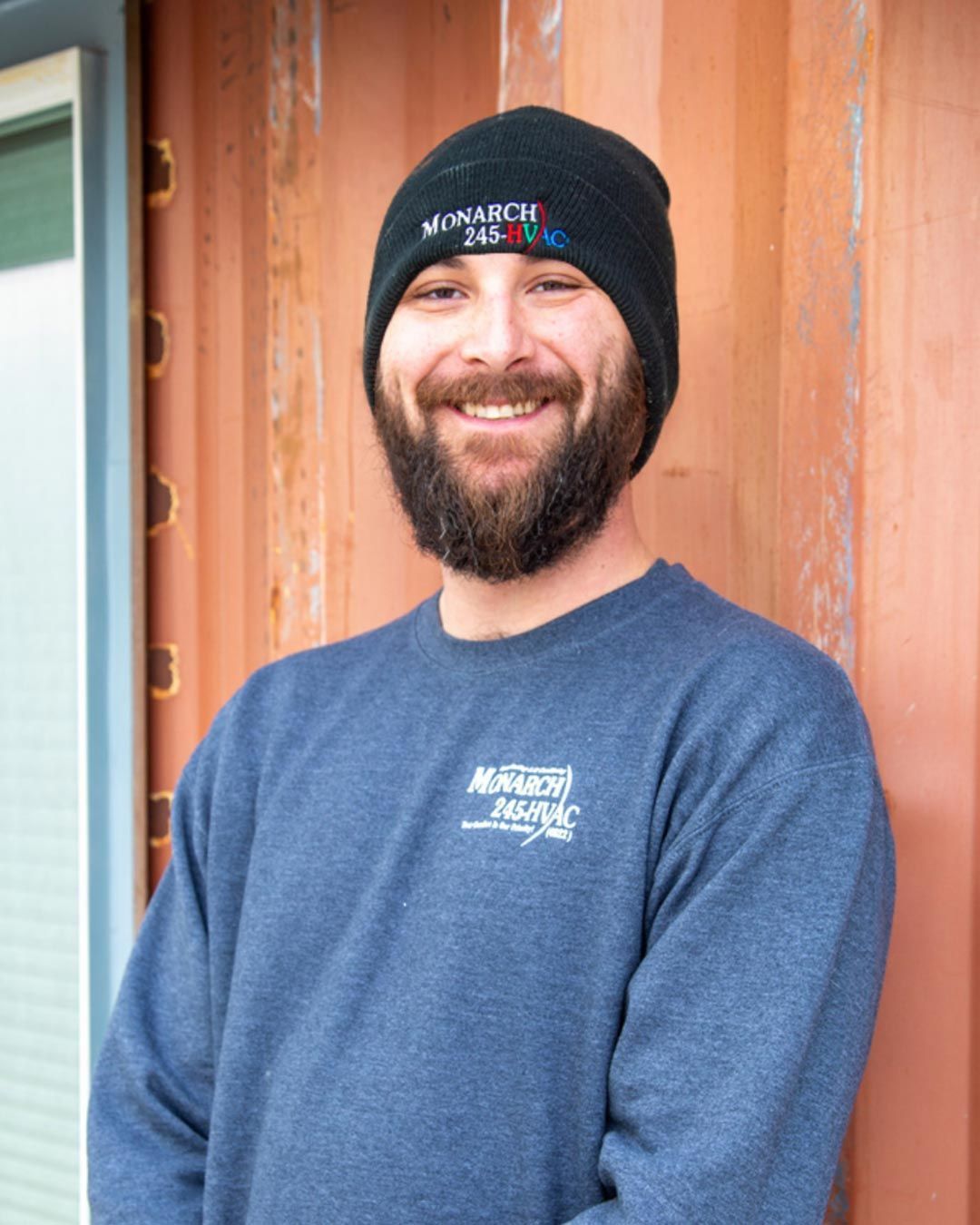 Man with a beard, wearing a beanie and sweatshirt, smiling in front of a rusty orange wall.