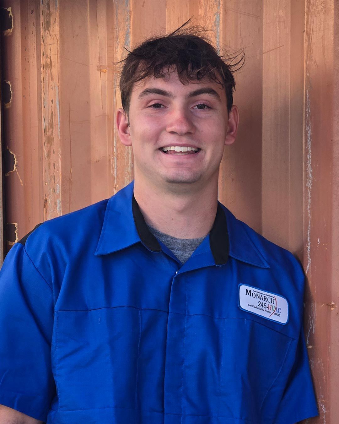 Smiling man in blue work shirt stands against a brown wall.
