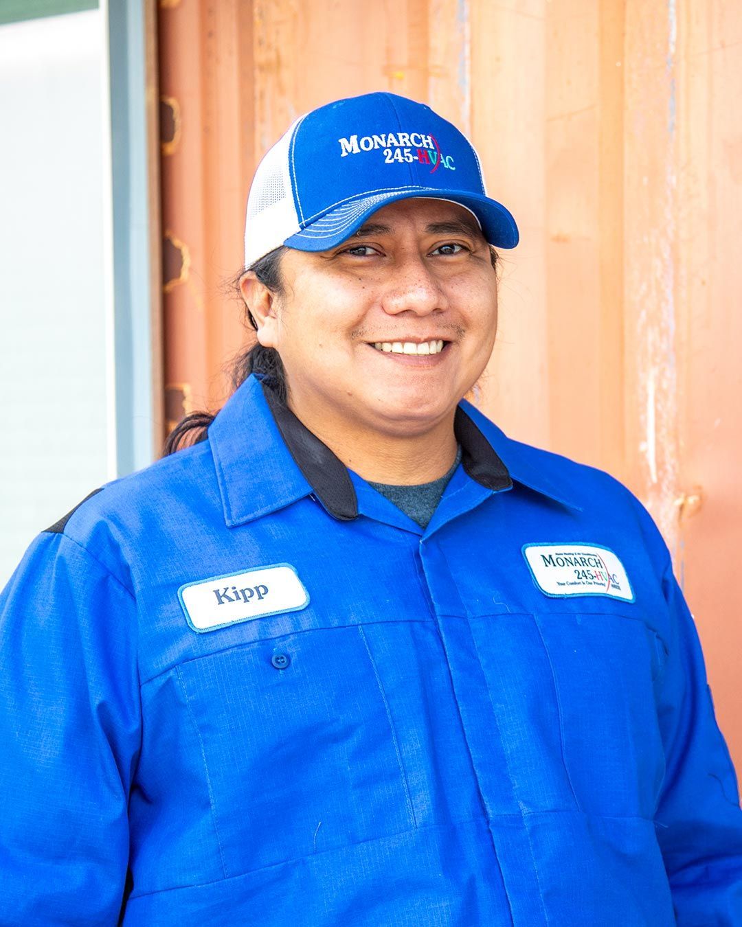 Man wearing blue uniform and cap, smiling. Name tag on chest, standing near a wall.