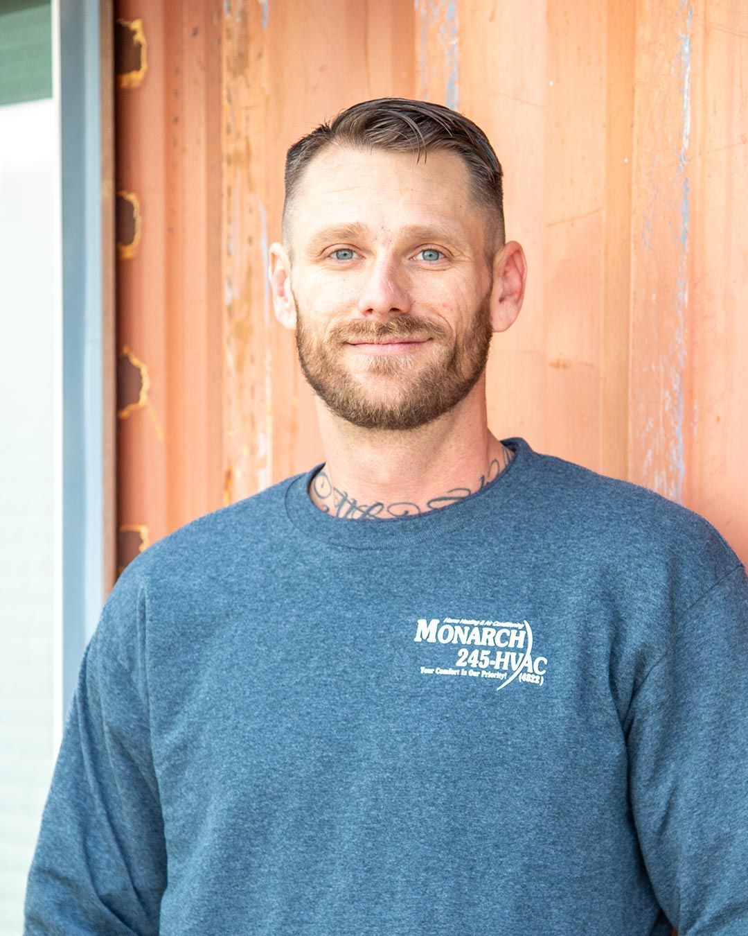 Man with a beard, in blue sweatshirt, standing next to a reddish-brown wall, smiling.