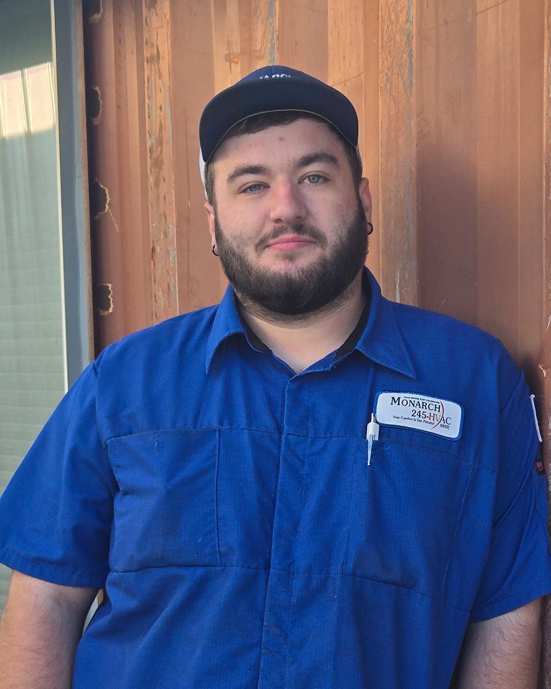 Man with beard wearing blue work shirt and cap, standing in front of a brown wall.
