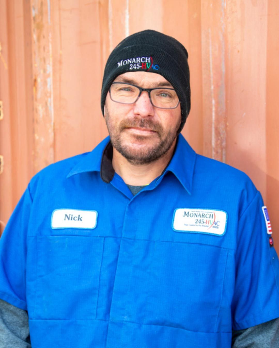 Man in blue work shirt and beanie, wearing glasses, standing in front of an orange background.