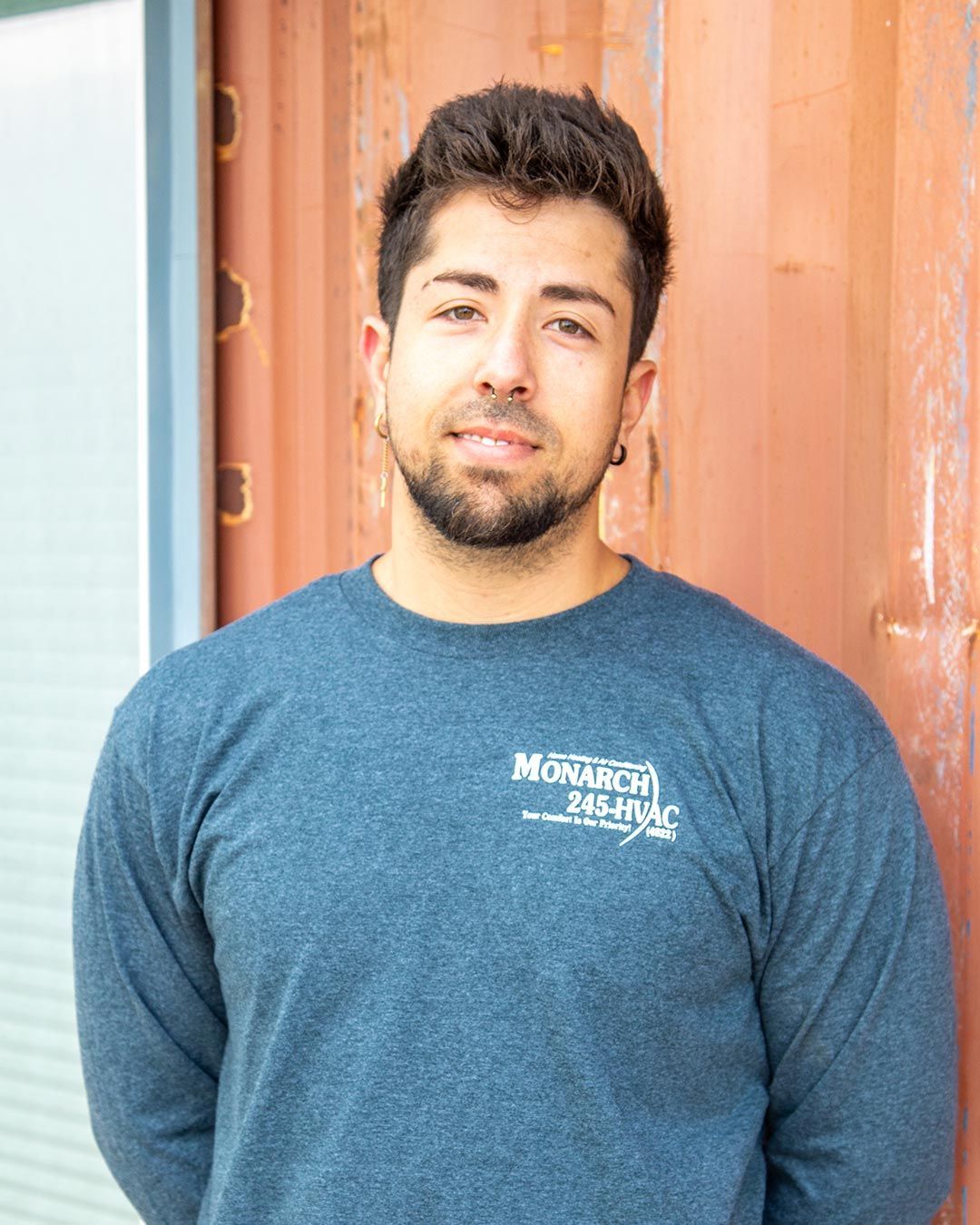 Man in blue long-sleeved shirt with logo, small beard, earring, standing in front of a rusty container.