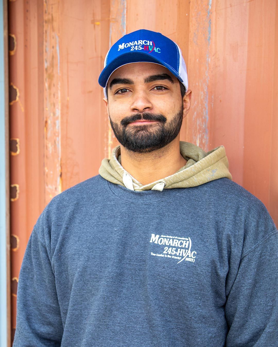 Man in blue cap and sweatshirt smiles, standing in front of a reddish-brown wall.