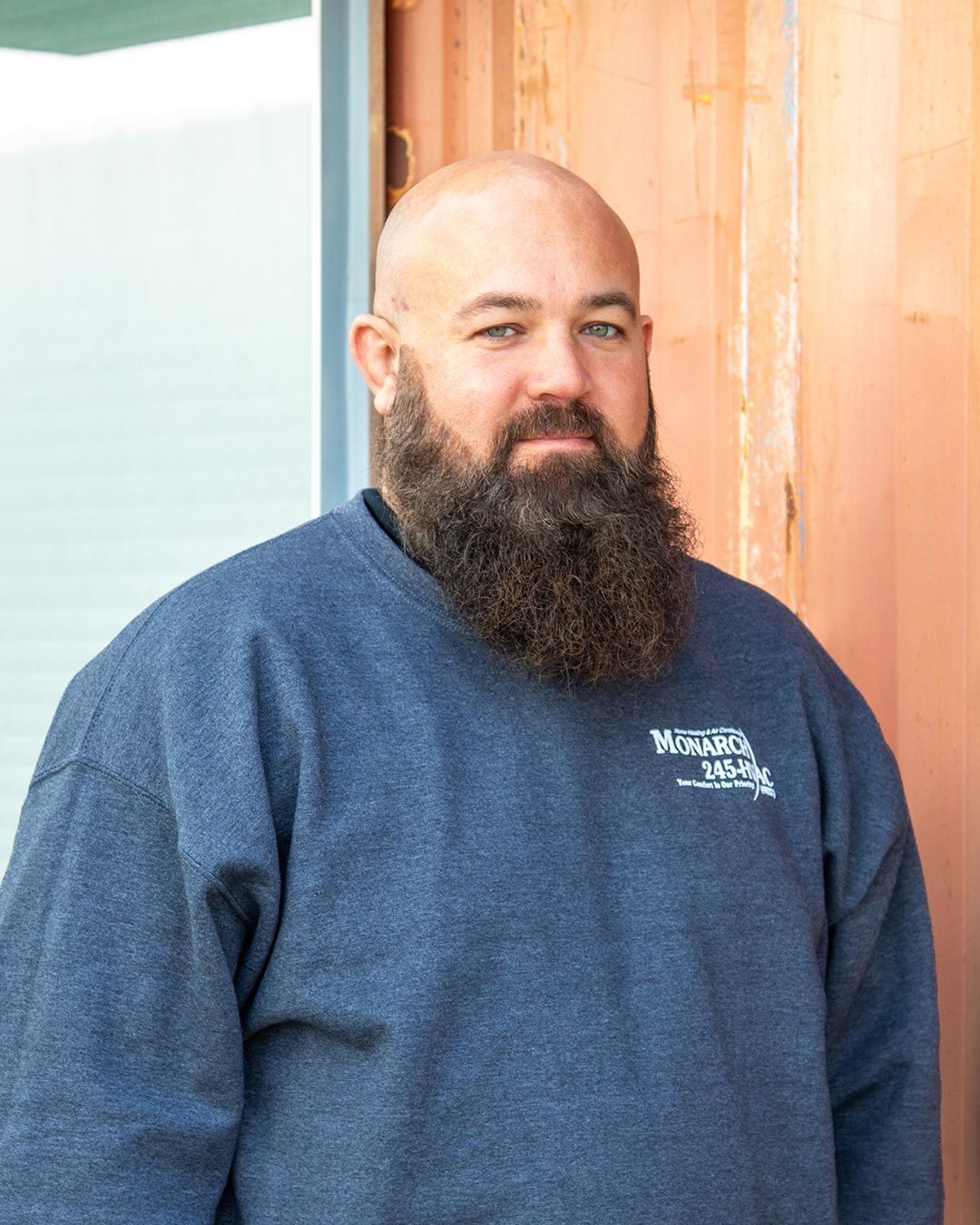 Bald man with a full beard wearing a blue sweatshirt with text, standing in front of a wood-paneled wall.