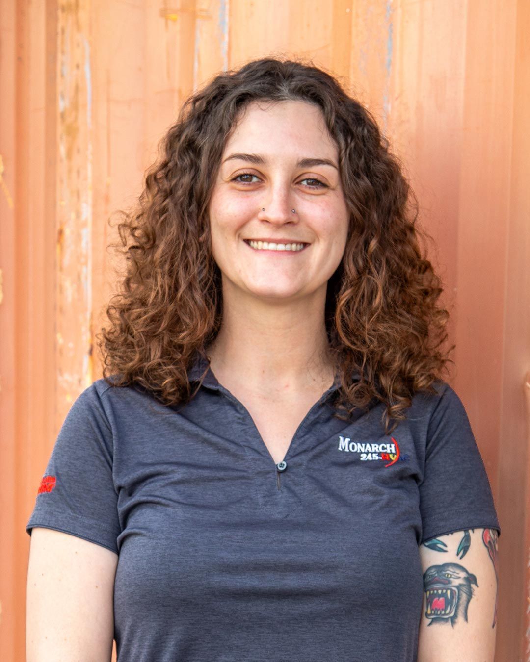 Woman with curly brown hair smiling, wearing a blue shirt with a company logo, in front of a rustic orange backdrop.