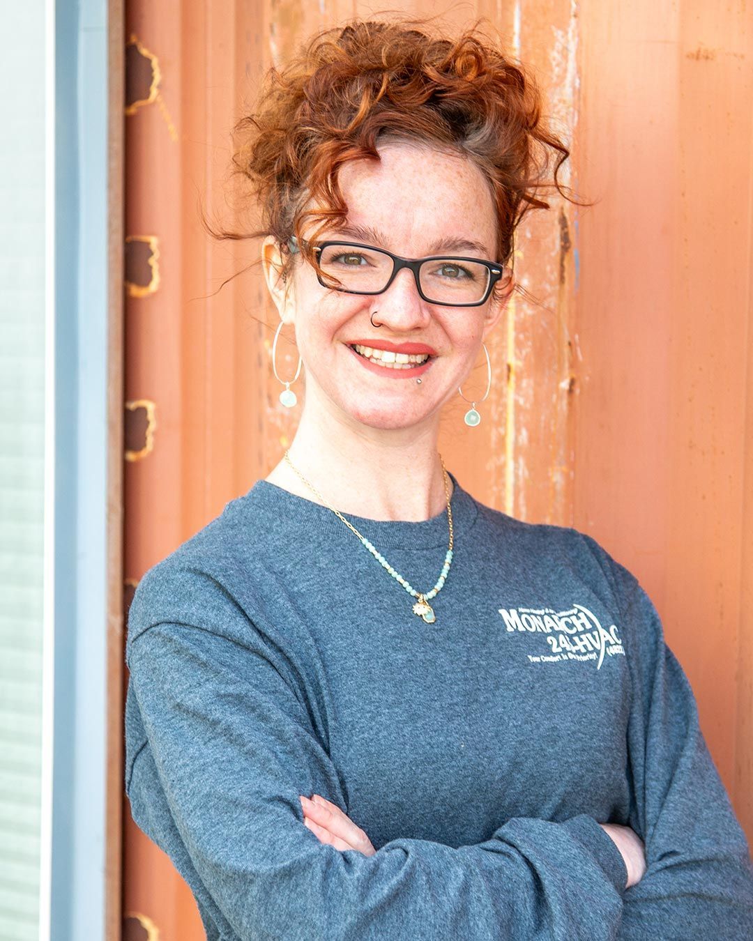 Woman with curly red hair, glasses, arms crossed, smiling. Leaning against rusty orange wall, wearing a grey shirt.
