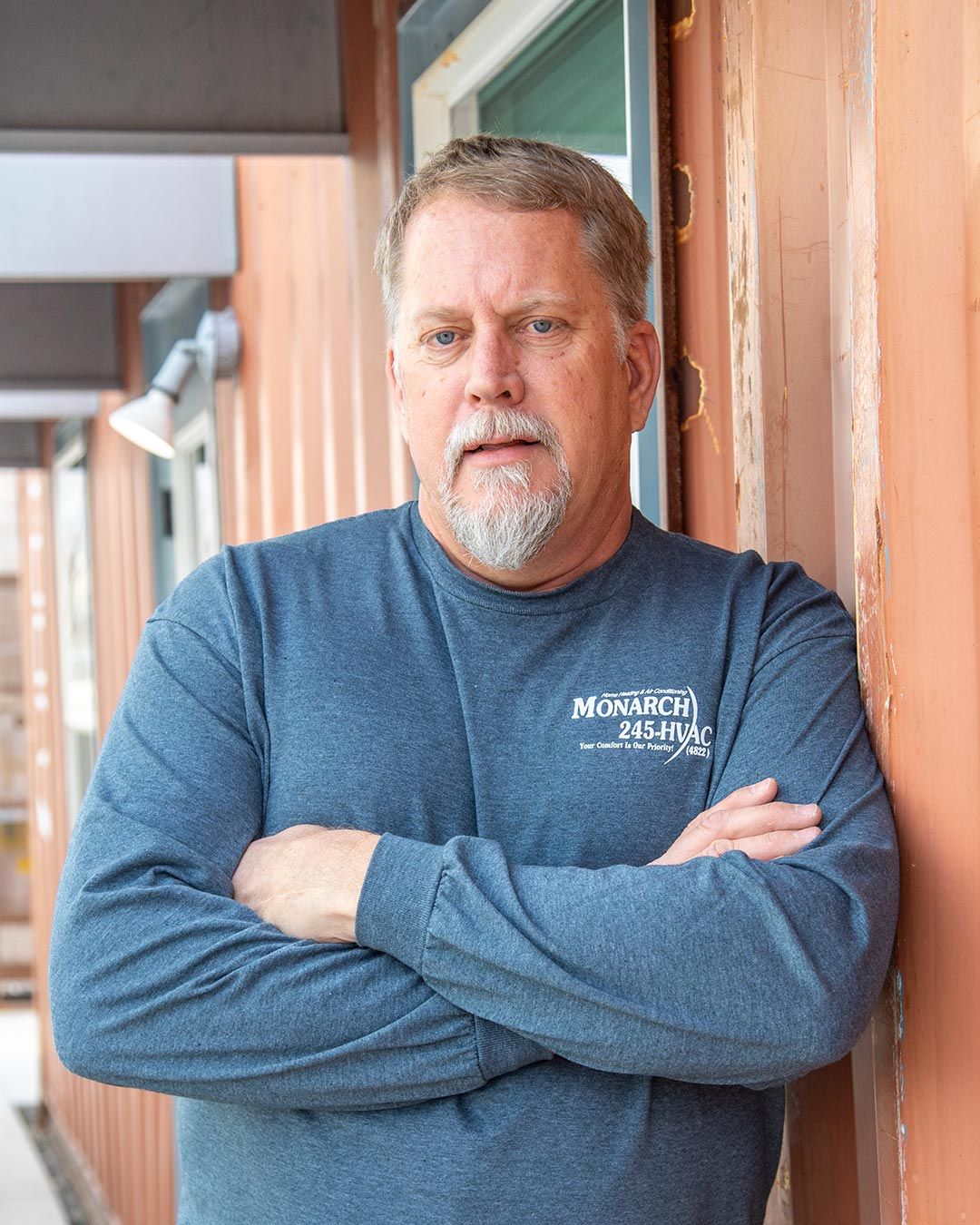 Man with arms crossed, leaning against a rusty orange wall, wearing a blue long-sleeved shirt with a logo.