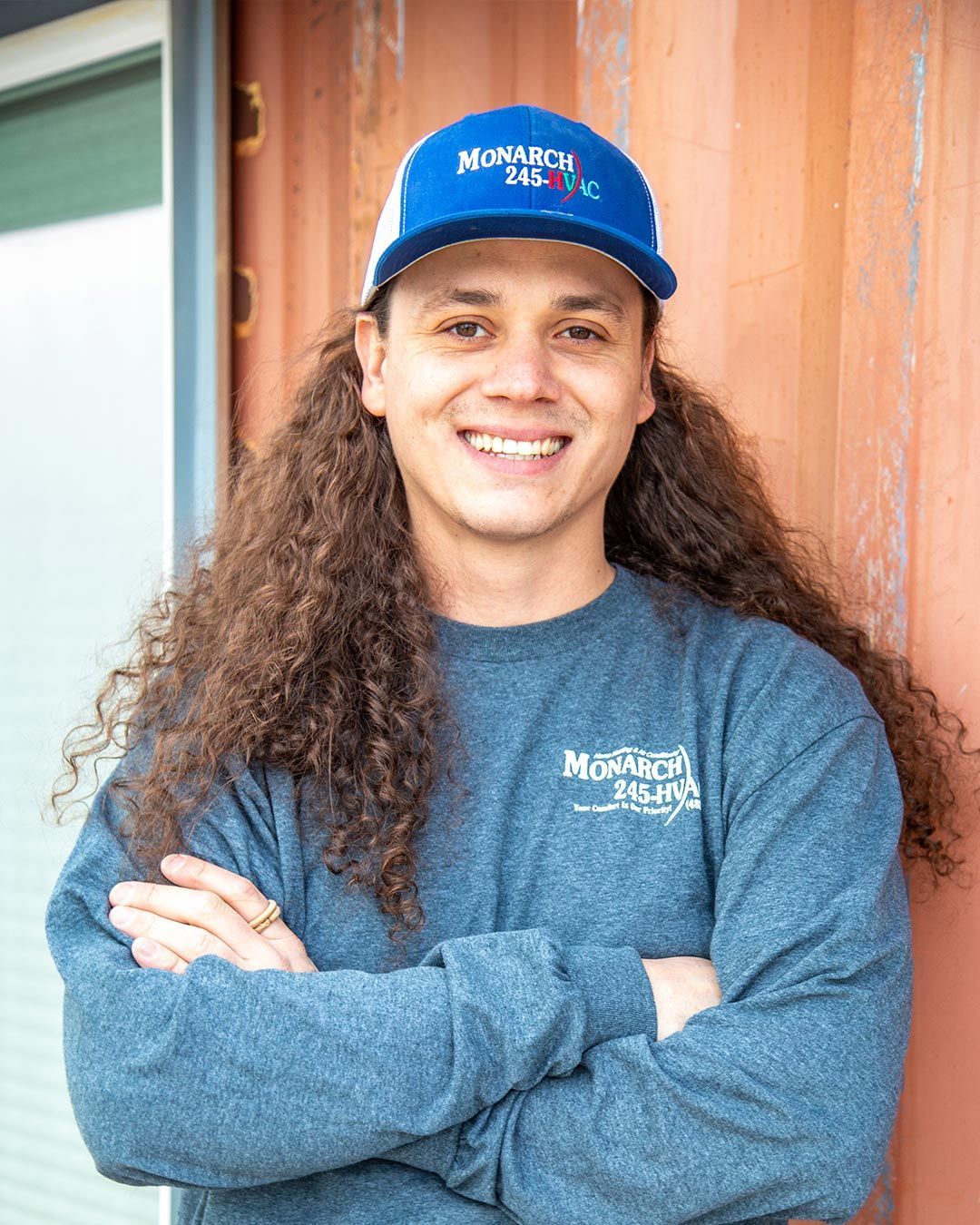 Man with long curly hair wearing a blue hat and shirt, arms crossed, smiling, leaning against a rusty container.
