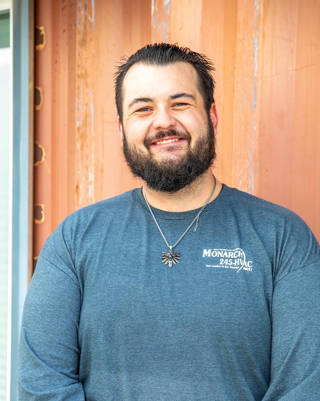 Man with dark hair and beard, smiling. Wearing a gray long-sleeved shirt, standing in front of an orange wall.
