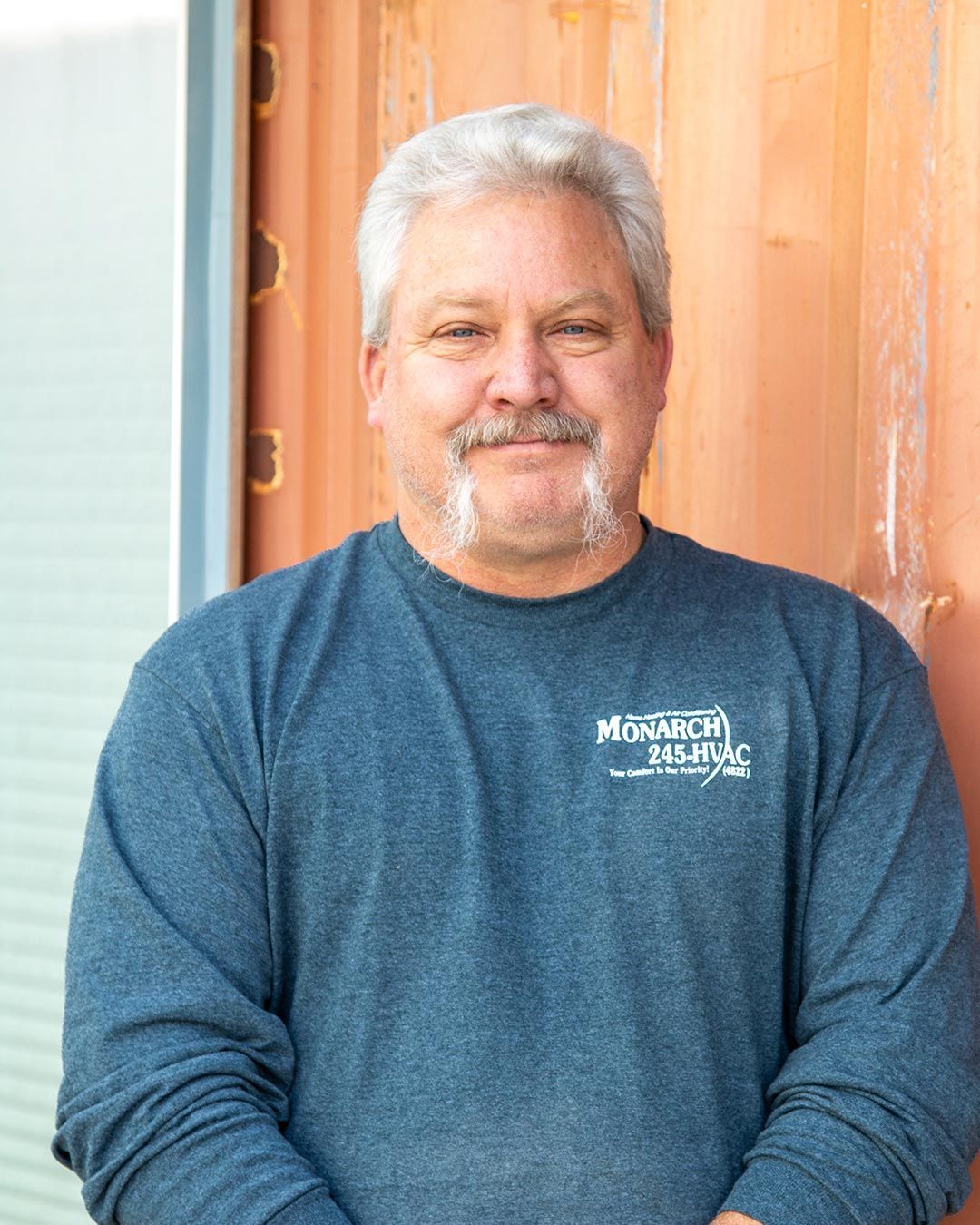 Man with gray hair and goatee in a blue long-sleeve shirt stands outside, smiling.