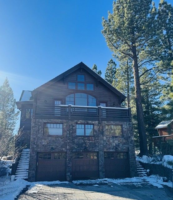 Snowy wooden cabin with a balcony and garage, surrounded by tall pine trees under a bright blue sky