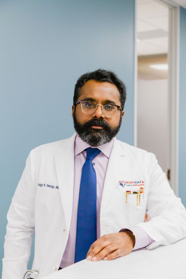 Doctor in white coat and blue tie standing in a medical office, facing the camera.