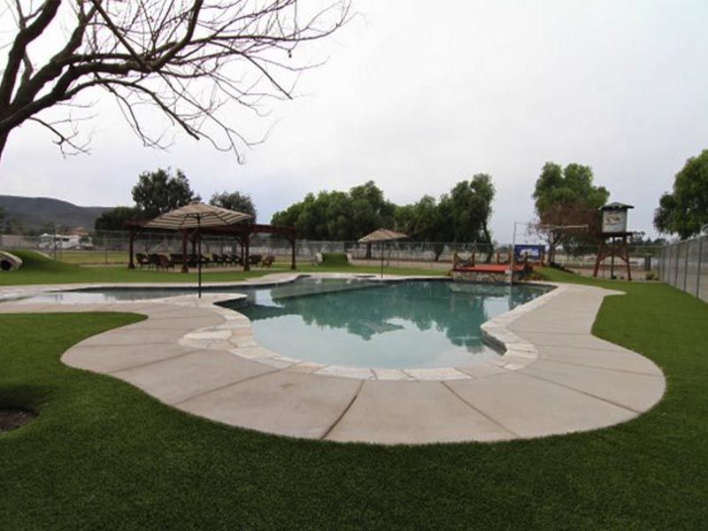 Swimming pool with curved stone deck in a grassy park, with gazebos and trees under an overcast sky