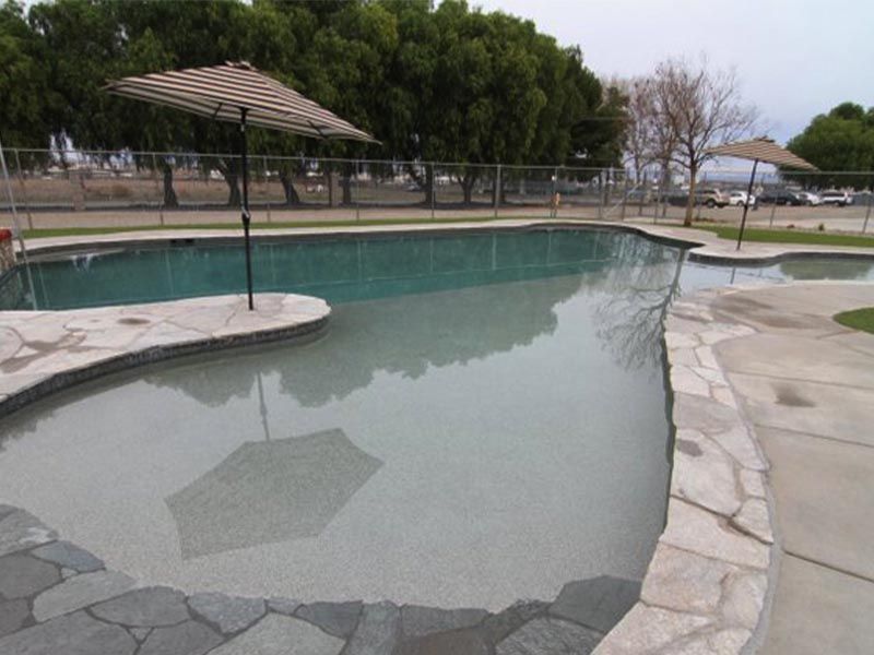 Empty outdoor swimming pool with pale green water, stone deck, and shaded patio umbrellas.