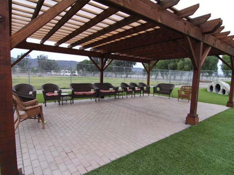 Covered outdoor patio with wooden pergola, wicker chairs, paved floor, and green lawn beside a lake.