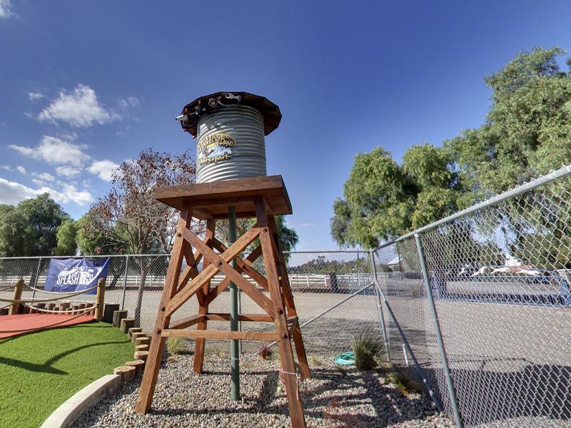 Wooden water tower on a playground beside a chain-link fence under a blue sky