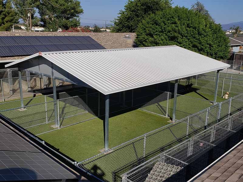 Covered metal pavilion with white roof on a fenced green sports court, viewed from above