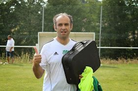 Un homme portant une chemise adidas blanche tient une valise noire et lève le pouce.