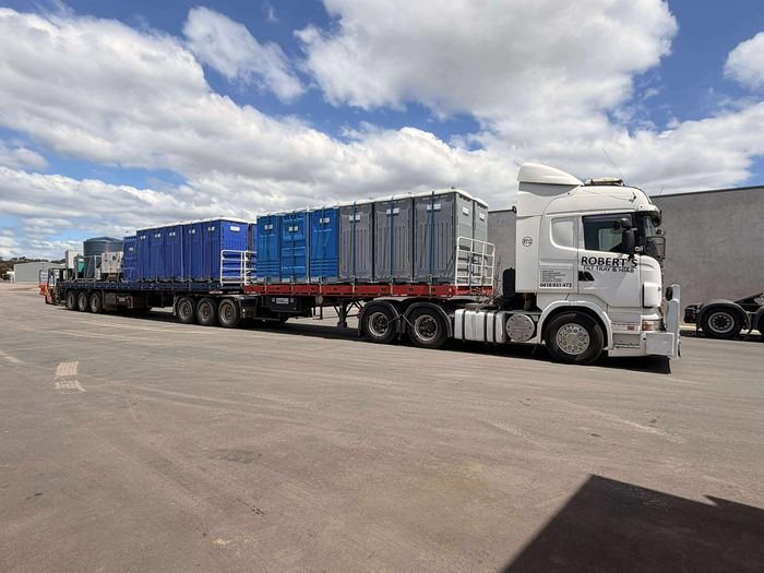 Long flatbed truck loaded with blue and gray shipping containers in a paved lot under a cloudy sky