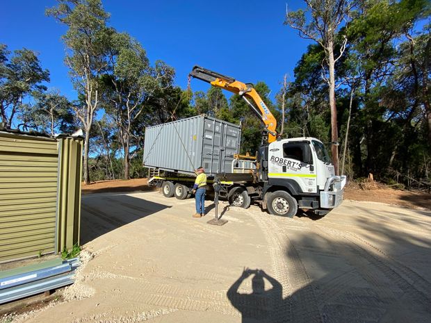 Crane truck unloading a gray shipping container beside a dirt road in a wooded area.
