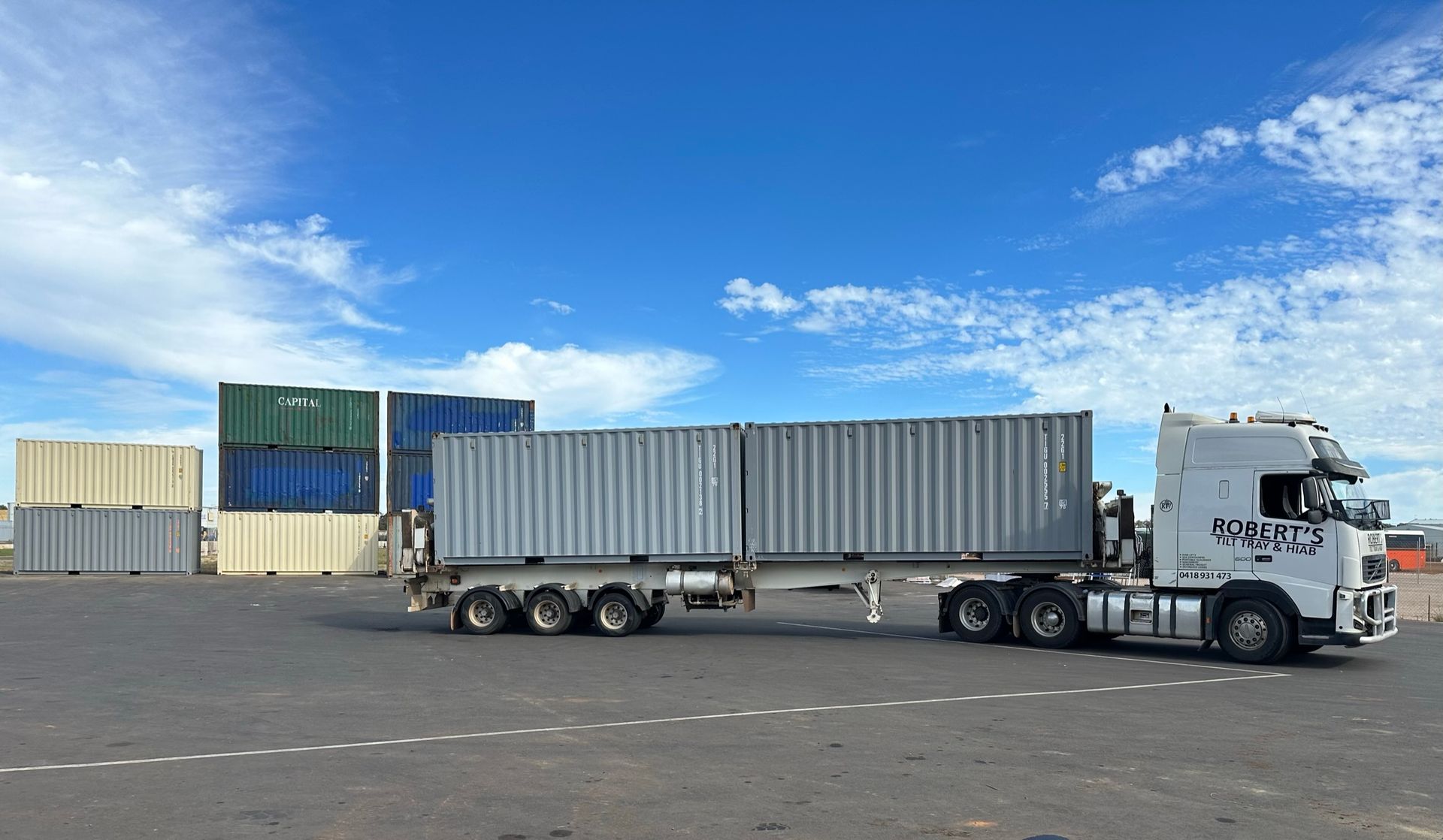 White semi-truck hauling a long gray shipping container in a port yard under a blue sky