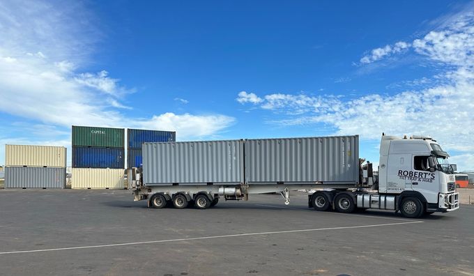 White semi-truck hauling a long gray shipping container in a port yard under a blue sky
