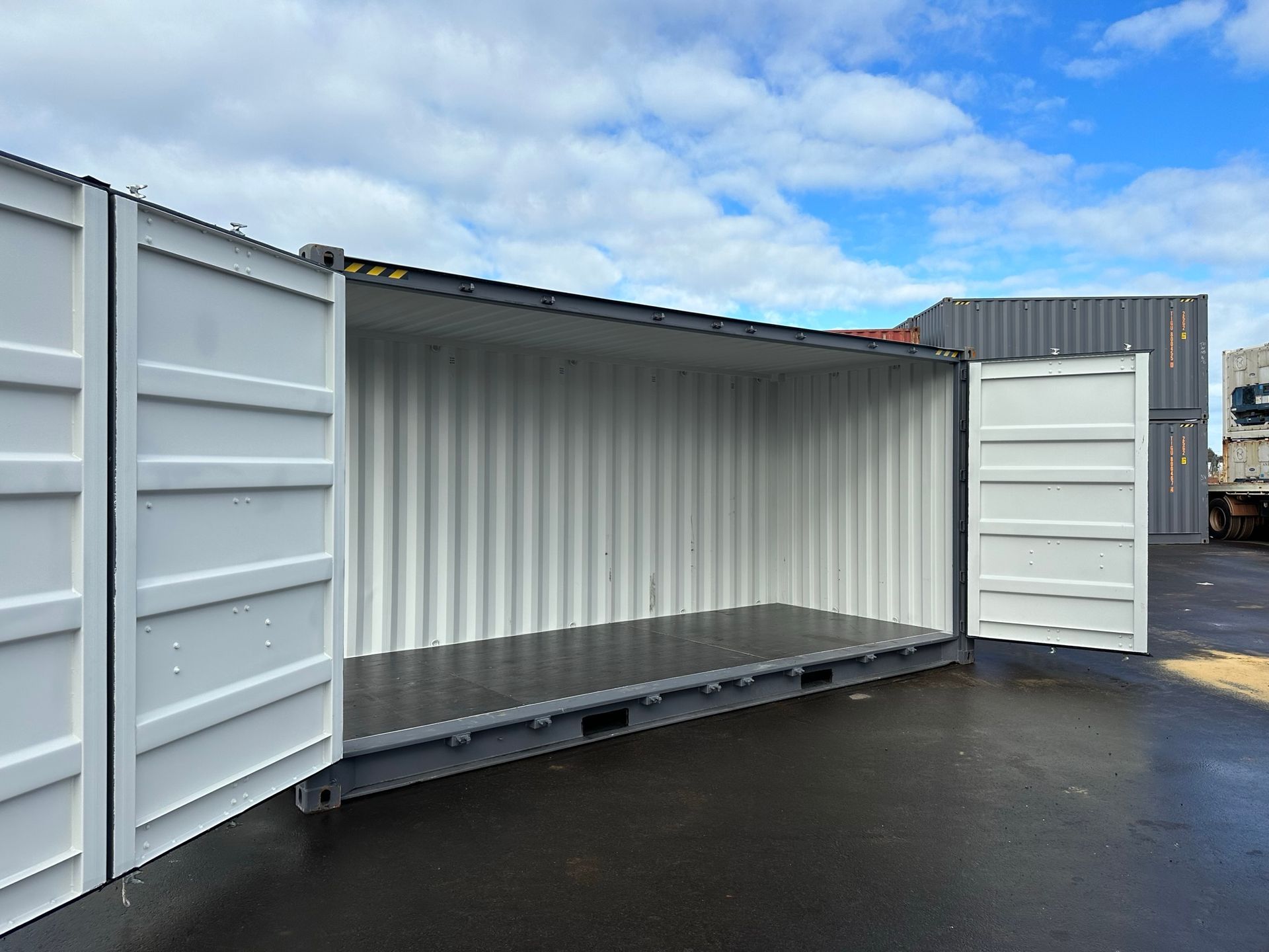 Open white shipping container with side doors on wet pavement under a cloudy sky