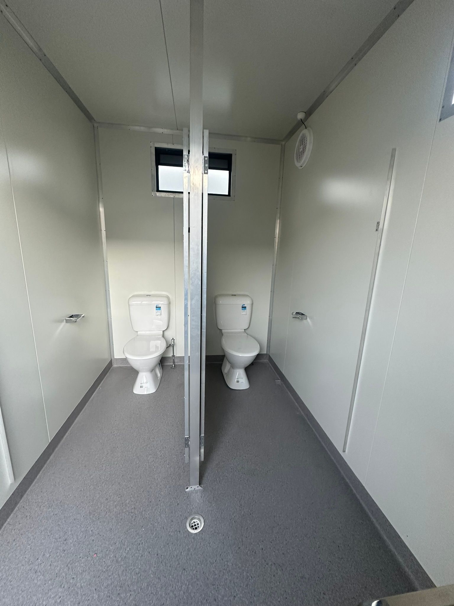Two toilet stalls facing a central aisle in a plain gray restroom trailer
