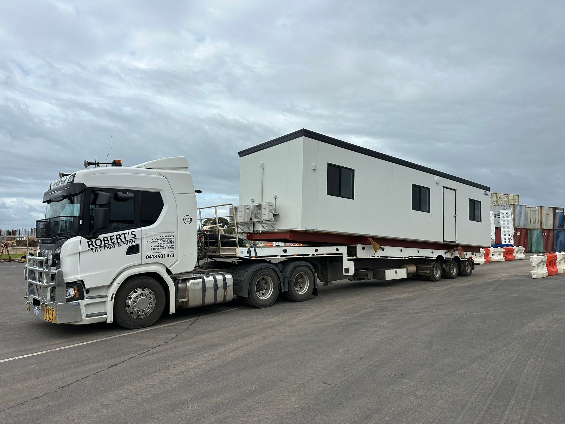 White semi-truck hauling a large modular building on a road under a cloudy sky