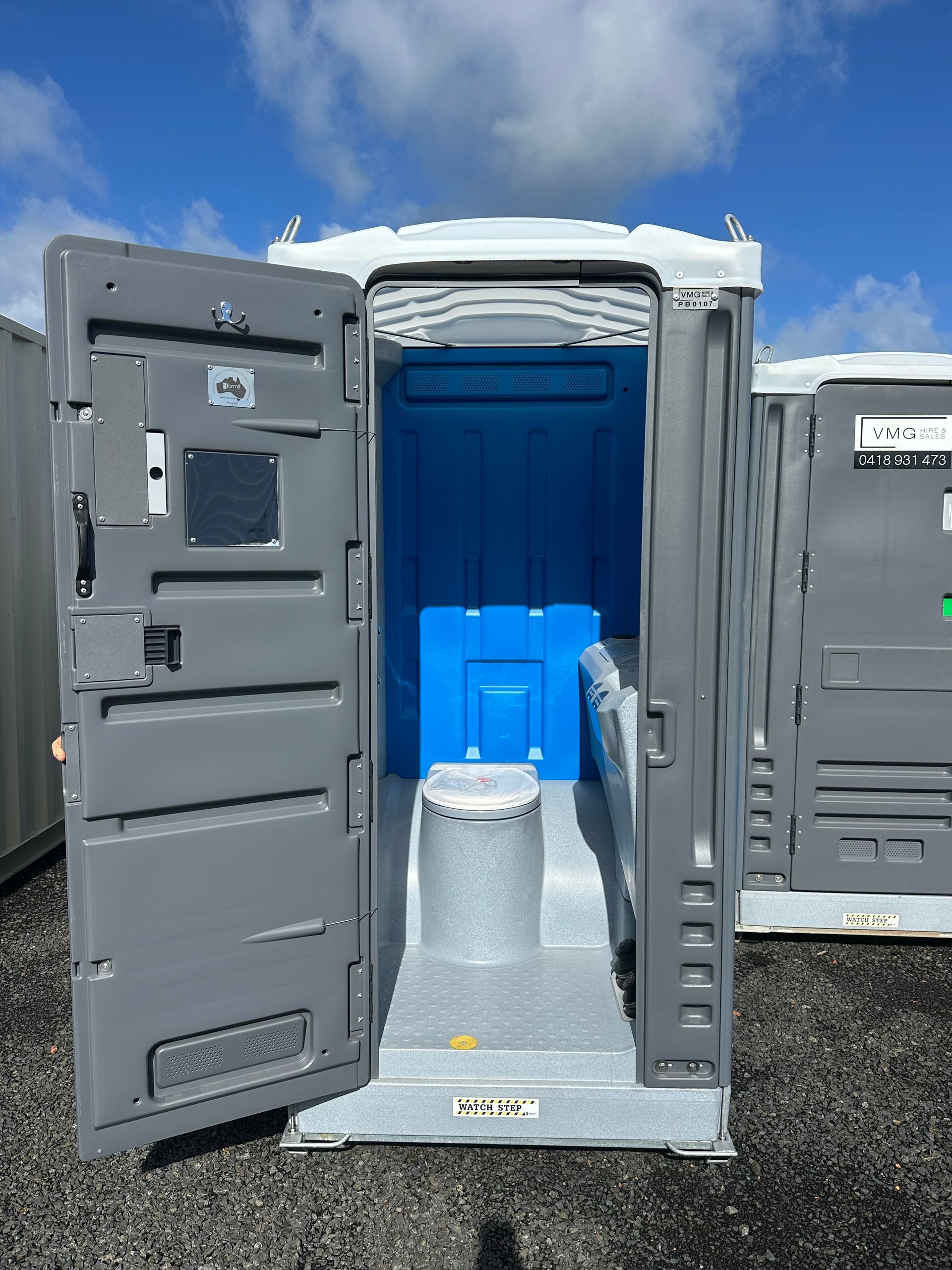 Open portable toilet with blue interior and white toilet seat outdoors under a sunny sky