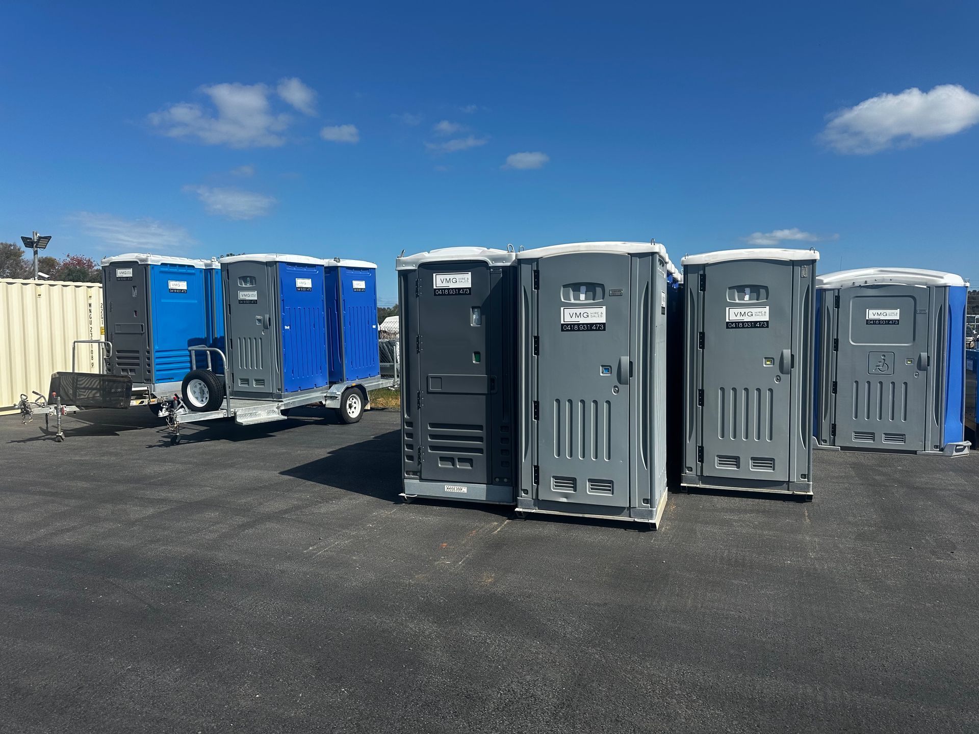 Portable toilets lined up on a paved lot under a blue sky, with blue and gray units.