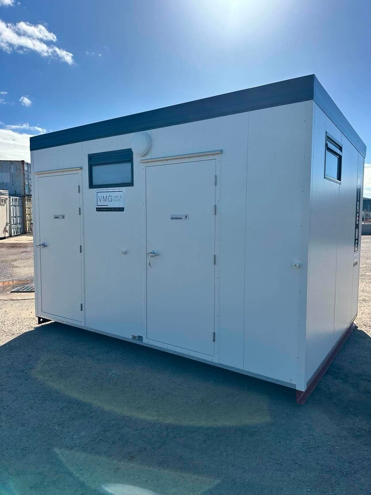 White modular utility cabin with a door and small window, sitting on a sunlit gravel lot.