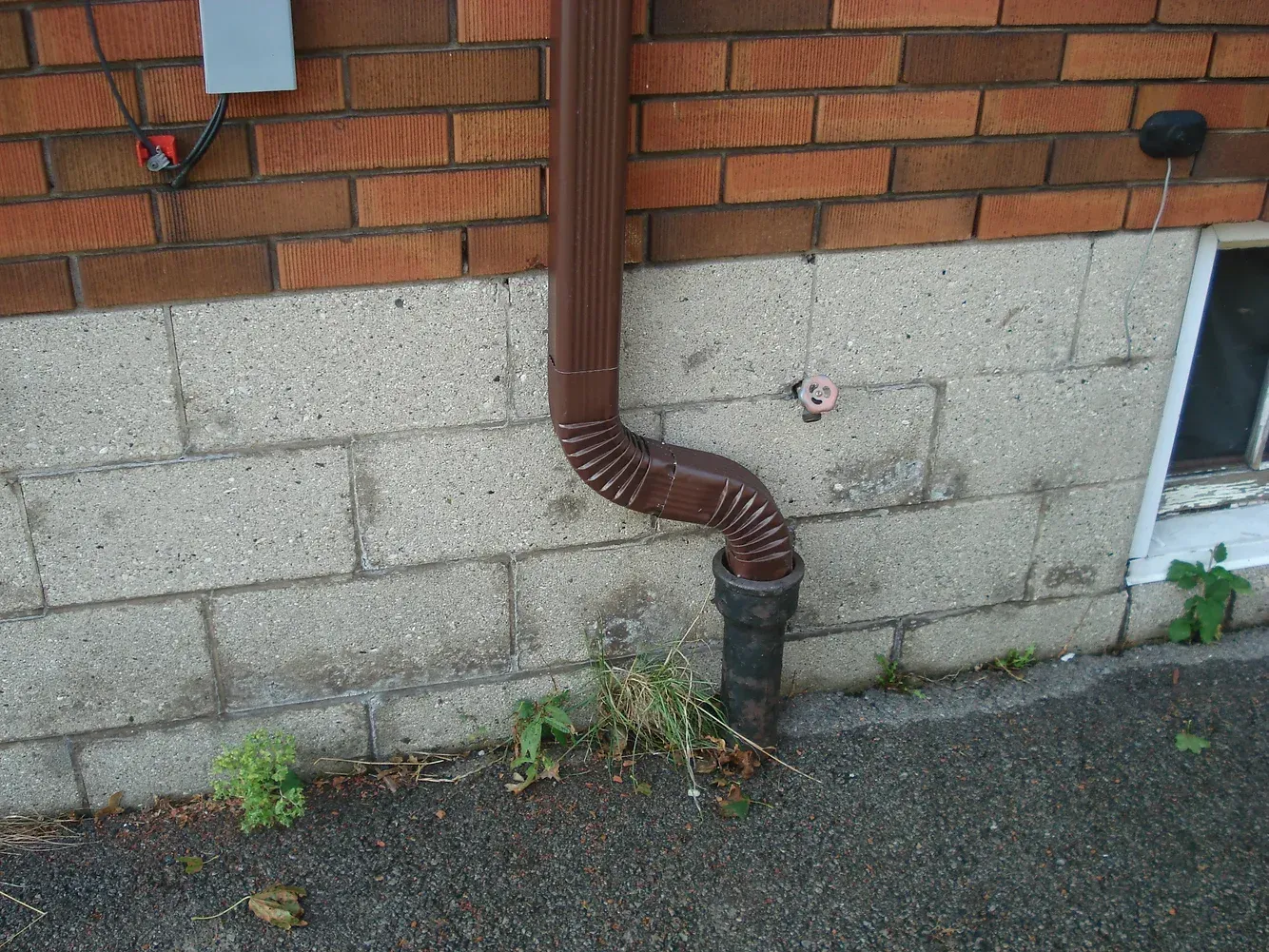 A brown downspout extends from a brick wall into a pipe in the ground against a concrete foundation.