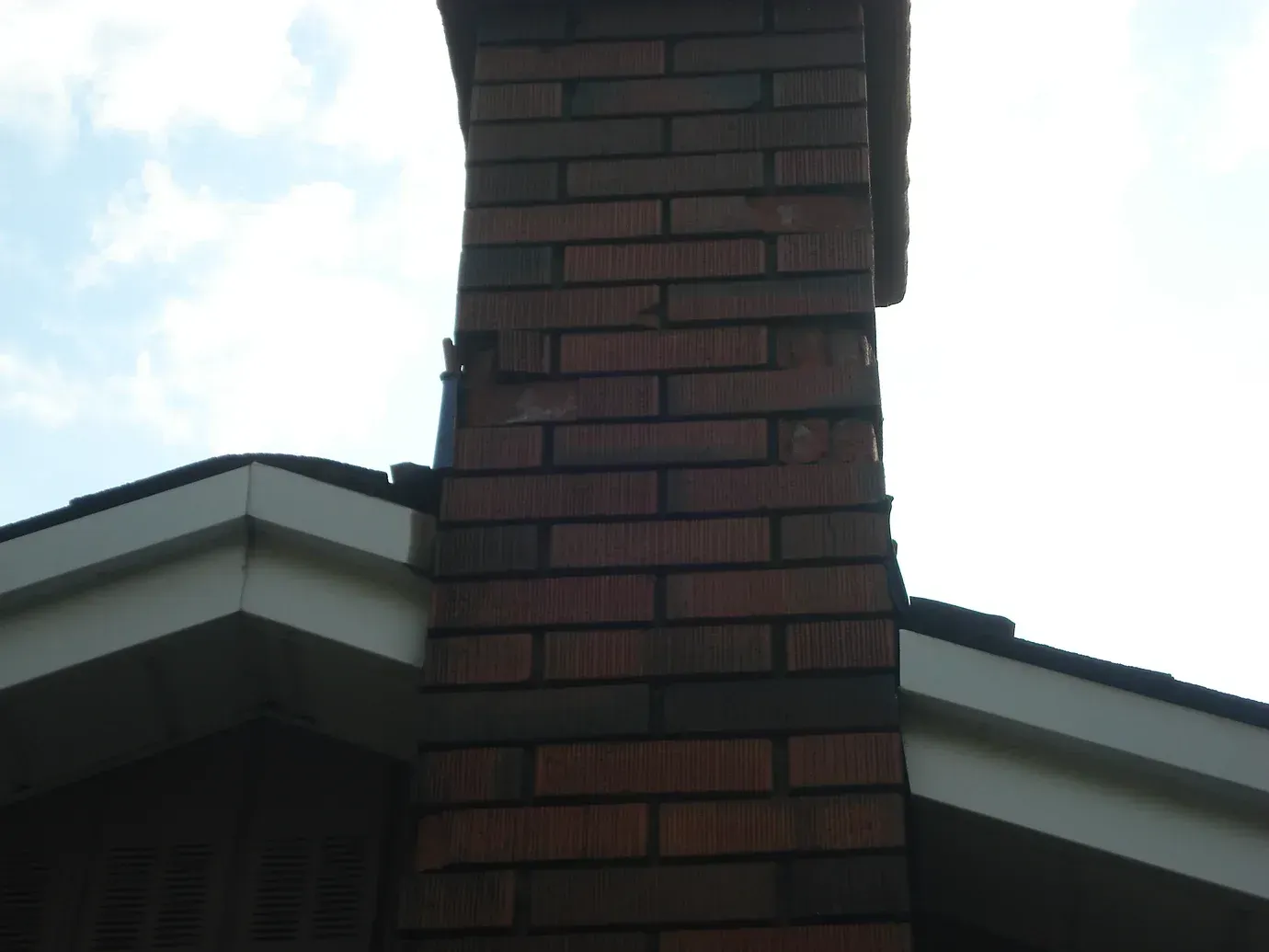 A brick chimney rising from the intersection of a house roof, showing signs of cracked mortar joints.
