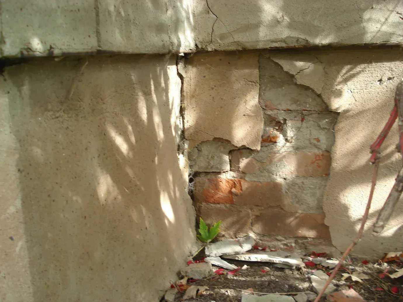 A corner of a foundation with crumbling, peeling concrete plaster, revealing red brick masonry underneath.