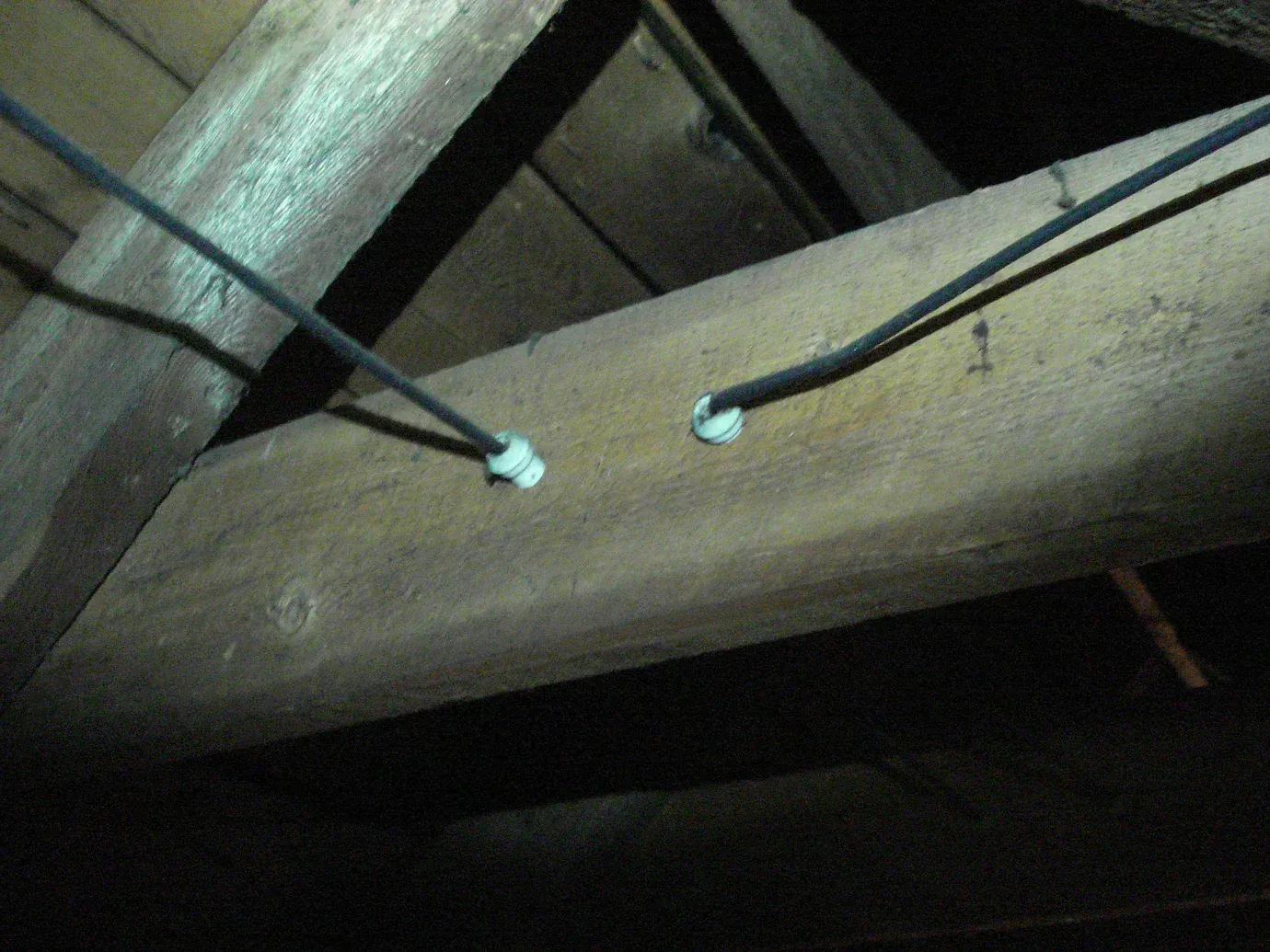 Two black electrical cables pass through white plastic insulators mounted into a wooden ceiling joist in an attic.