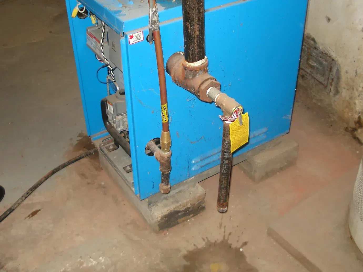 A blue boiler leaking water onto a concrete floor from a discharge pipe connected to a side fitting.