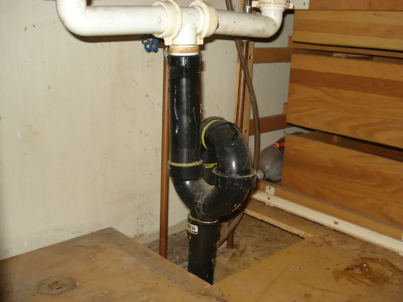 A black S-trap plumbing pipe connected to white sink drain pipes under a wooden cabinet.