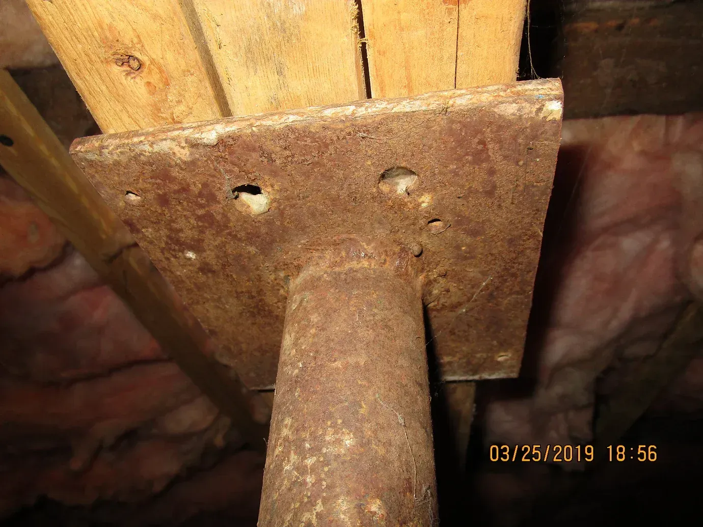 A rusty metal support post with a square mounting plate attached to a wooden beam in a crawlspace or basement.