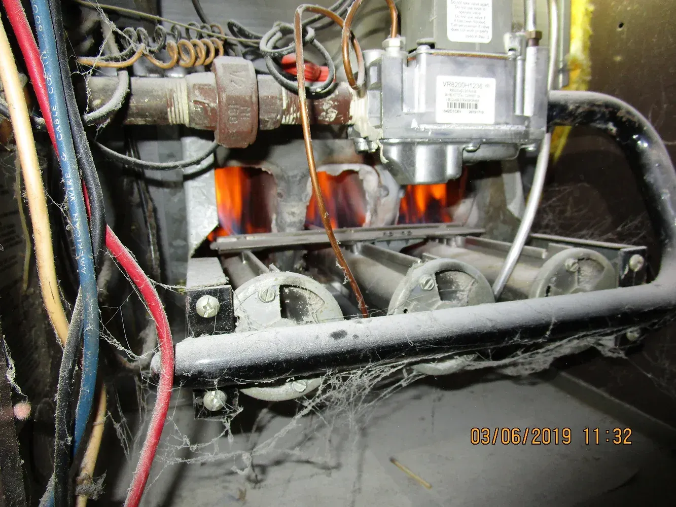 Interior of a furnace showing lit burners, gas valve, wiring, and heavy dust accumulation on the components.
