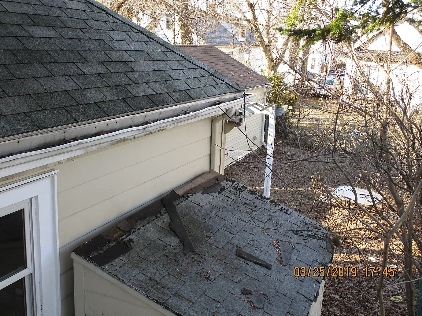 Exterior of a house showing a detached gutter and damaged shingles on a lower roof section near bare trees.