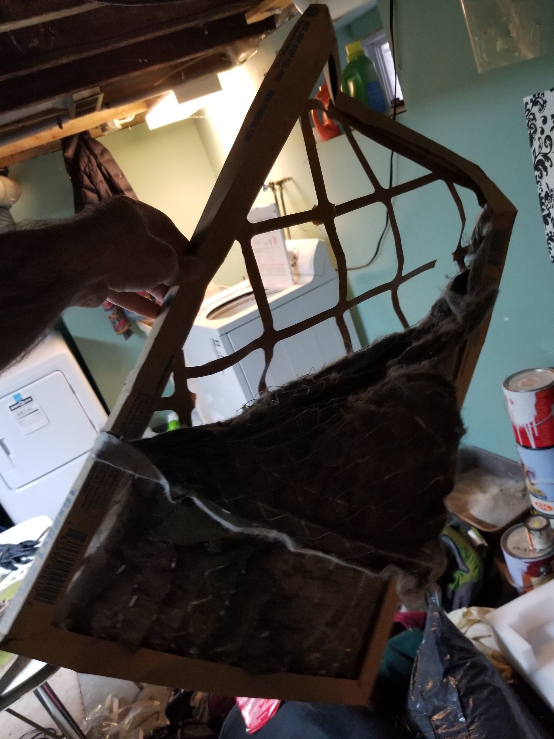 A person holds a dirty, wooden, lattice-style window frame with torn screen mesh in a cluttered room.