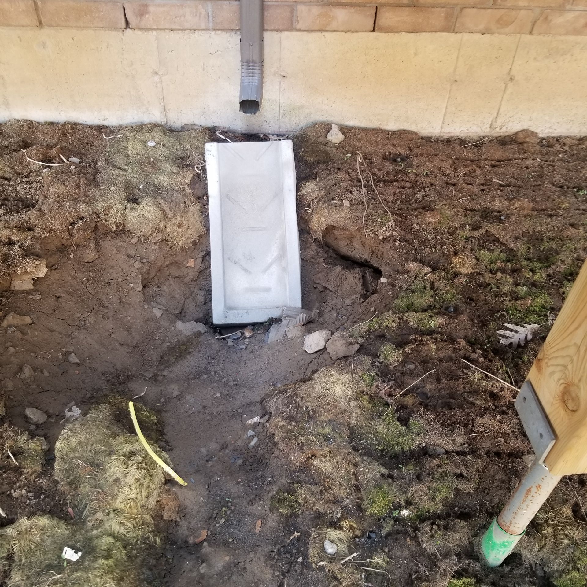 A metal downspout extender directs water onto a concrete splash block set in dirt beside a brick foundation.