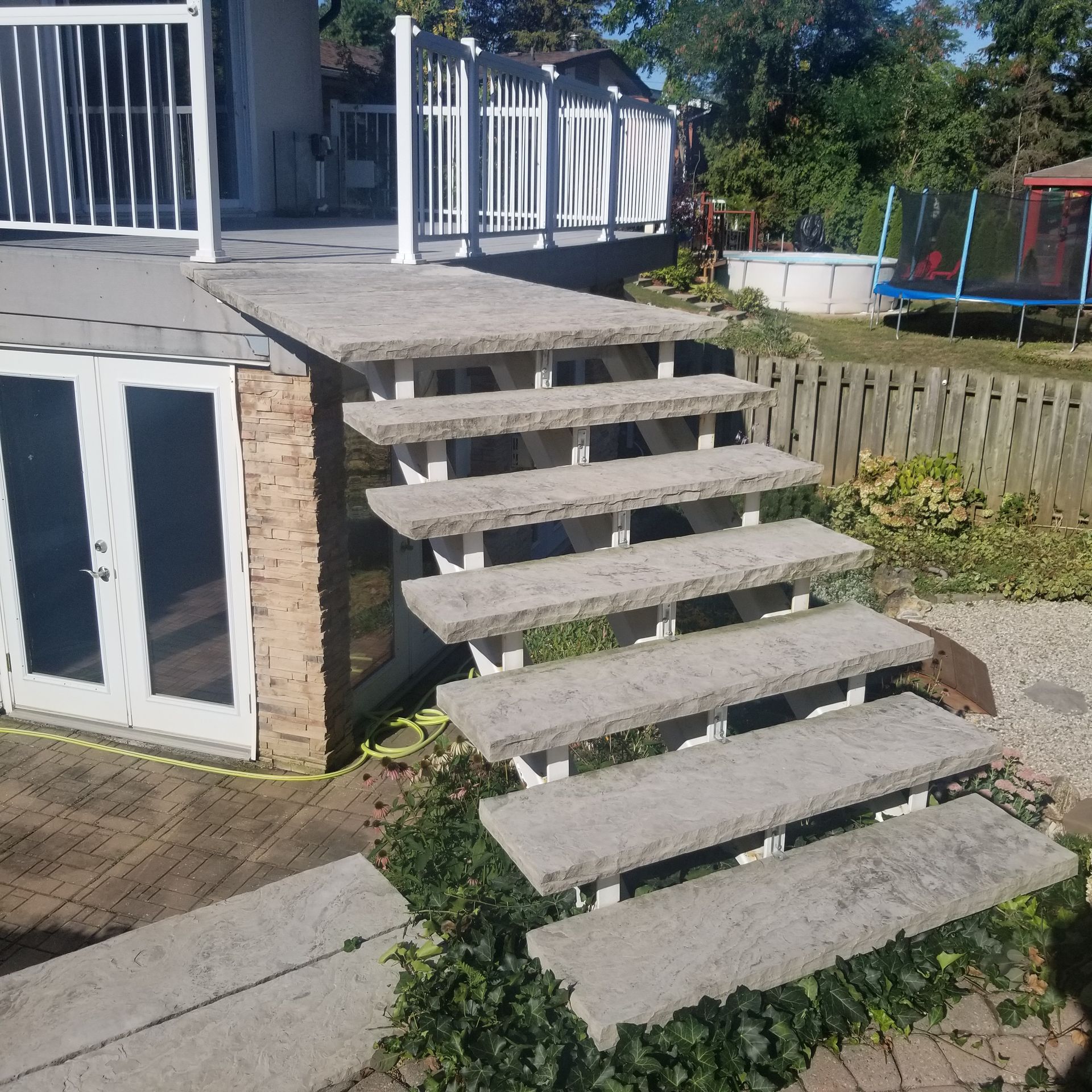 Stone stairs with a white railing lead from a backyard patio to an elevated wooden deck next to a house.