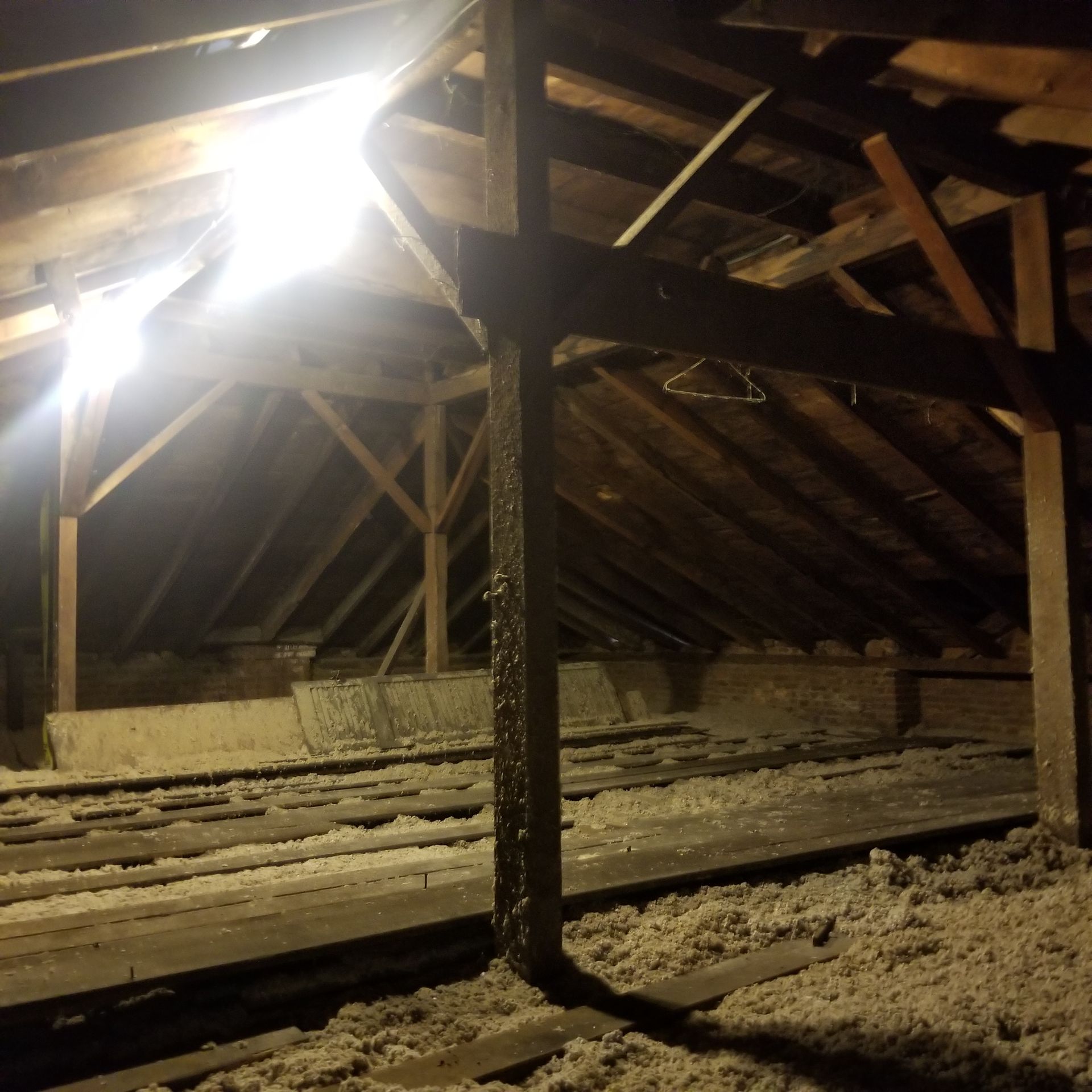 A dimly lit wooden attic with exposed rafters, insulation between floor joists, and a bright light source on the left.