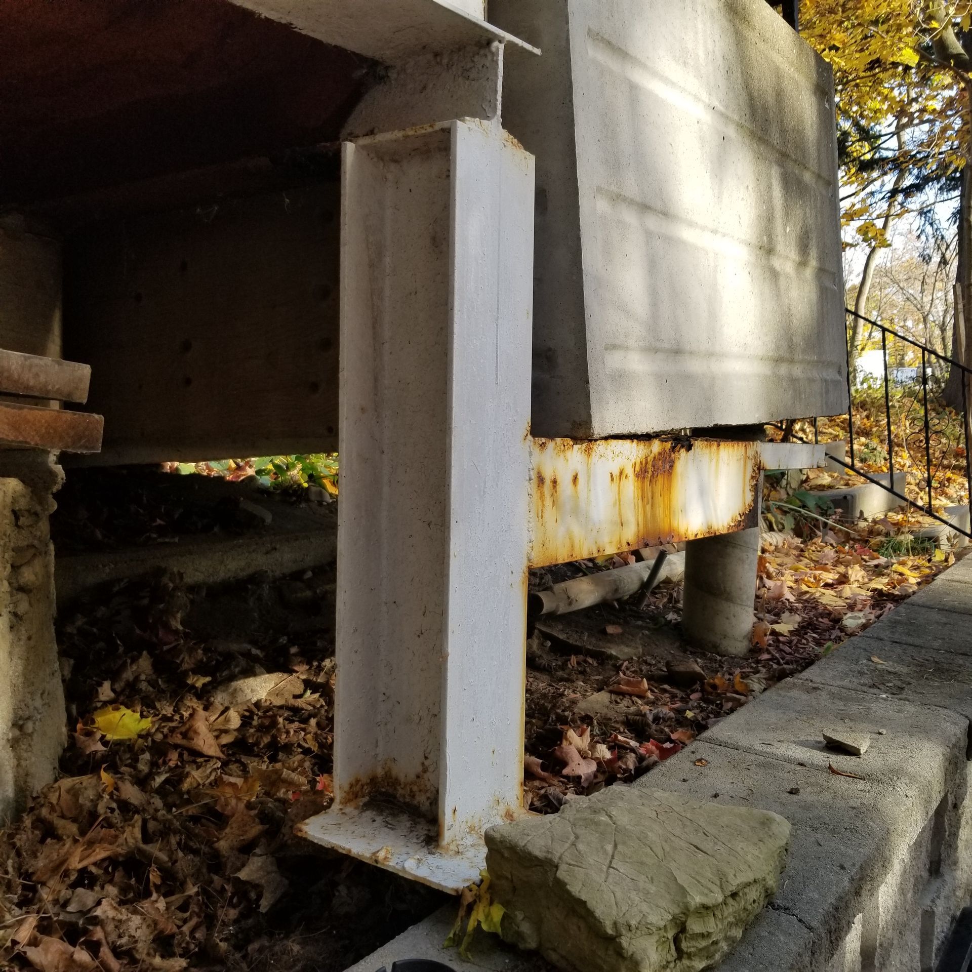 A steel support column and rusted beam holding up a concrete structure over an area covered in dry fall leaves.