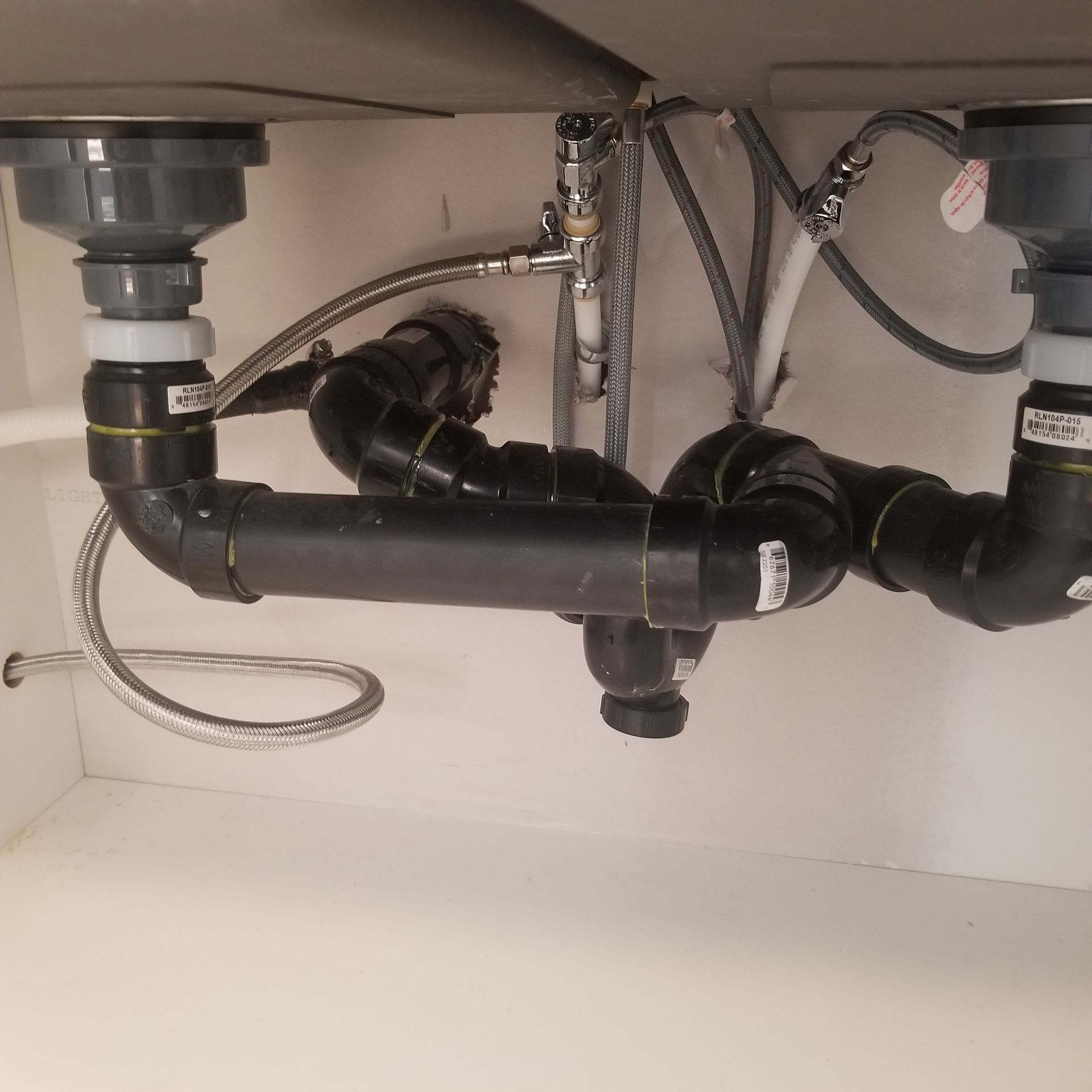 A view under a kitchen sink showing black PVC pipes connected to a double basin drain, with water supply lines behind.